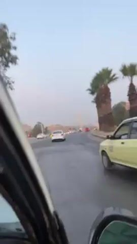 The view from a moving car is shown driving on an urban road, lined with tall palm trees. Other cars are visible driving in the same direction, and buildings are seen in the distance. The sky is a pale blue.