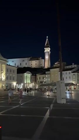 The video shows a night scene in a town square. A tall clock tower with illuminated windows and a clock face stands prominently in the background. Buildings with illuminated windows surround the square. Several people are walking in the square, and some are seated at tables under covered outdoor seating areas. The ground is paved, and shadows are cast by the buildings and lights. A tall, slender pole, possibly a flagpole, is visible in the foreground. The sky is dark, indicating it is nighttime.
