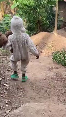 The video shows a young child walking outdoors. The child is wearing a hooded white knit jumpsuit and green sandals, and is carrying a brown teddy bear on their left shoulder. They walk uphill along a dirt path, pause to look at the camera, then continue walking towards a tree and building. The environment includes trees, plants, and a simple building in the background.