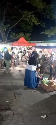 The video shows a night market with stalls selling various goods, possibly clothing or accessories, displayed on the ground and on tables. Several people are seen walking around and browsing the merchandise. Some individuals are wearing hijabs and traditional Islamic clothing. The market appears to be set up outdoors under trees, with artificial lighting illuminating the scene. The overall atmosphere suggests a busy and lively marketplace.