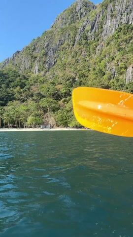 The video captures a first-person perspective from a kayak moving through clear, green-tinted water towards a white sand beach. A steep, rocky mountain covered in dense green vegetation dominates the backdrop. The sky is a clear, bright blue. The movement of the kayak is achieved with an orange paddle, frequently dipping into the water. Another rocky island can be seen to the right. The video gives an impression of calm and clear weather.