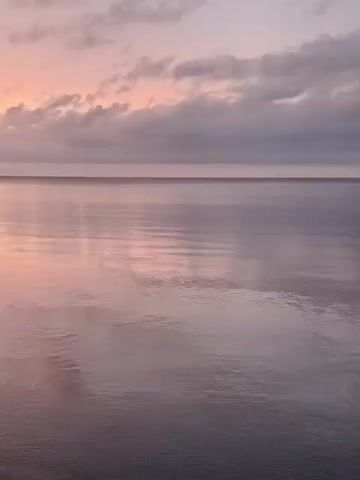 The video shows a serene body of water at sunset. The sky is painted with hues of pink, orange, and purple clouds, reflecting beautifully on the calm water's surface. A dark object, possibly a log or debris, floats in the middle of the water on the left side of the frame. The camera pans slightly to the right, revealing more of the colorful sky and the gentle lapping of waves against a sandy shore on the bottom left. The overall atmosphere is tranquil and visually stunning.