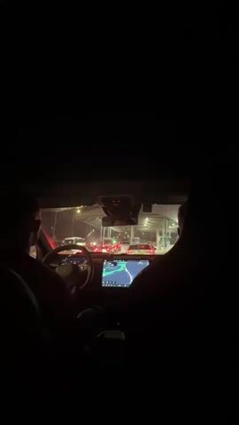 The video, shot from the interior of a vehicle at night, shows a driver and passenger facing forward, looking out at a traffic queue at a border crossing. The driver's hands are on the steering wheel, and the car's navigation screen is visible, displaying a map with a route. The border crossing area is illuminated by artificial lights, with multiple vehicles lined up. The scene is dark, with the interior of the car mostly silhouetted.