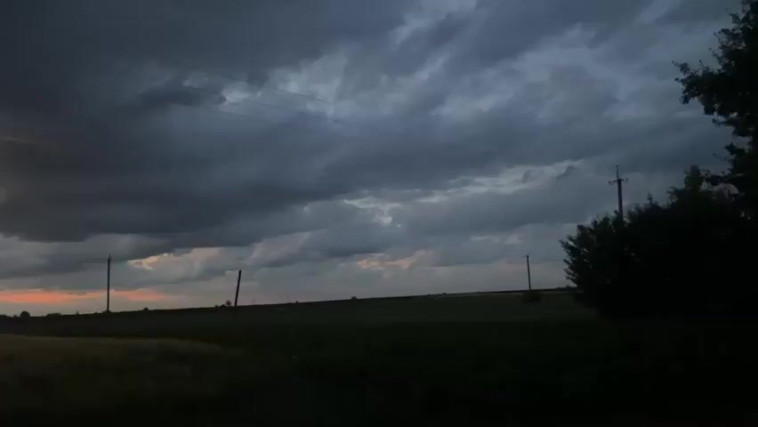 The video shows a wide shot of a rural landscape at dusk. The sky is filled with dark, dramatic clouds, with hints of orange and pink from the setting sun peeking through on the left side of the frame. In the foreground, there are fields of crops, appearing dark and silhouetted against the sky. Several utility poles with wires strung between them are visible across the landscape, as is a silhouetted tree on the right side of the frame. The overall mood is somber and somewhat ominous, suggesting an approaching storm.
