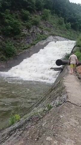 The video shows a man standing on a concrete embankment next to a spillway where a large volume of water is flowing rapidly down a steep concrete channel into a reservoir or river below. The man is wearing a light-colored t-shirt and shorts. He is looking at the flowing water. The spillway is covered in white foam and churning water. The surrounding environment is a hillside covered in green trees and rocks. There is a black pipe extending from the side of the spillway. The camera pans slightly to the left showing more of the churning water and the embankment.