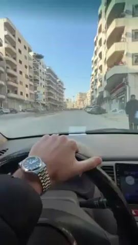 The scene is a first-person perspective from inside a car driving down a city street. The driver's left hand, adorned with a silver watch, rests on the steering wheel. Buildings line both sides of the street, varying in height and color, predominantly beige. Cars are parked along the curbs. The sky is clear and blue, suggesting daytime. The car's dashboard is visible, featuring an infotainment screen displaying time and temperature. The overall atmosphere suggests an urban environment with signs of wear and tear on the buildings.