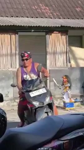 The video depicts a Balinese Barong statue standing on a pedestal in front of a building. The statue is white and gold, with a red face, long black hair, and a sword in its hand. People walk past the statue, including a woman in a purple shirt and sarong. A man is visible on a motorcycle. The building in the background has a corrugated metal roof and wood-framed windows.