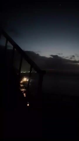 The video shows a dark, grainy shot of a twilight sky over a body of water. The sky transitions from a deep blue at the top to a faint orange at the horizon. There are scattered clouds. In the foreground, a dark railing or barrier is visible on the left side, partially obscuring the view. Lights from a distant town or settlement can be seen on the left, near the water's edge. There is also a bright spot, possibly a star or planet, visible in the upper part of the sky. The overall impression is that of a dimly lit coastal scene at dusk or dawn.