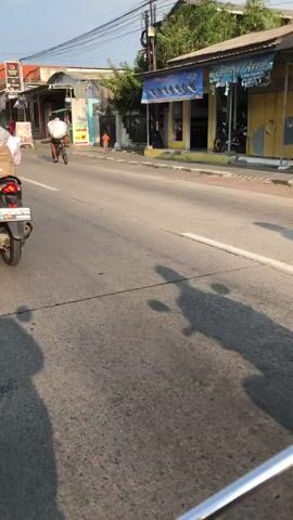 The video captures a street scene in Boro, East Java, during what appears to be the late afternoon or early evening, given the long shadows. The sun is low in the sky, casting a warm glow on the environment. The weather seems fair and clear.

The primary subjects are people engaged in everyday activities. In the foreground, a person is riding a motorcycle, their legs and feet visible, wearing casual clothing and sandals. The motorcycle itself is black and red. Further down the road, another person is cycling, carrying a large, round, white object on their head. They are wearing a light-colored shirt and shorts, and a backpack. In the background, a black SUV is driving on the road. A small child in an orange shirt is also visible, standing near the side of the road.

The scene is a typical street in a populated area, likely a commercial or residential district. Shops with awnings and signage line the right side of the road. One shop has a yellow facade and displays what appear to be shoes or bags in its window. Another shop sign is partially visible with Indonesian text. The road is paved and marked with lane lines. Vegetation, including trees, is present in the background.

The activity captured is daily commuting and transport. The motorcyclist and the cyclist are navigating the road, while the SUV is also part of the traffic. The overall impression is one of a lively, functional street with people going about their daily routines. No strong emotions are evident; the mood is neutral and observational.

There is some visible text on the shop signs, but it is largely illegible or too small to be definitively read. One sign on the yellow building appears to have a logo and text. A sign on a pole in the background is also present but unreadable. The context of Boro, East Java, provides a specific geographical reference for the scene.