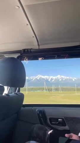 A POV shot from inside a vehicle shows a snow-capped mountain range in the distance under a clear blue sky. The foreground is dominated by the interior of the vehicle, including the back of a headrest and part of a car door. In the middle ground, a green field stretches towards the mountains, with several tall utility poles and some distant buildings. The view suggests a journey through a scenic, rural landscape.