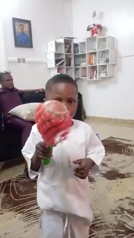 A young boy stands indoors, holding a red feather duster with a green handle. He is dressed in a white tunic and trousers. He turns the duster quickly in his hand. An adult male sits in the background on a black couch. There is a photograph on the wall behind the couch and a decorative shelf unit with white cube-shaped shelves. A bottle of water is on the floor. The room has light-colored walls and patterned carpet.
