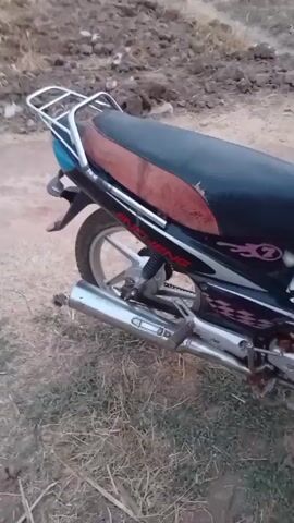 The video shows a motorcycle in a dirt and grassy area. The motorcycle is black and silver, with a torn, dark red and black seat. It has INCHENG written on its side, along with a "7" and a flame design. The motorcycle has a silver luggage rack. The camera moves around the motorcycle, showing the front wheel and handlebars. There are some books and papers placed on the flat area between the seat and handlebars. A voice states that the bike is for sale and provides contact information.