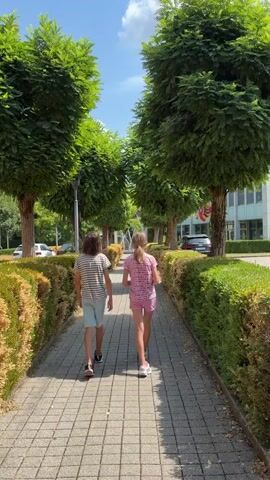 Two children, a boy and a girl, walk away from the camera down a paved pathway lined with trimmed bushes and trees. The boy, on the left, wears a striped t-shirt and light-colored shorts. The girl, on the right, wears a pink t-shirt and pink shorts. The pathway is made of square, gray pavers arranged in a checkerboard pattern. The bushes on either side are green on the right and brown and dry on the left. Behind the bushes on the right is a building with windows, and flags are visible on poles in front of it. Cars are parked on the left side of the pathway, beyond the bushes. The sky is blue with scattered white clouds. The trees are tall and leafy, forming a canopy over the pathway. The scene appears to be outdoors on a sunny day.