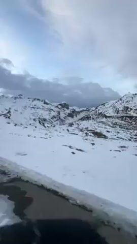 The video shows a first-person perspective of someone riding an e-bike on an asphalt road through a snowy mountain pass. The road is mostly clear, with patches of snow and ice, especially along the edges. Snow-covered mountains rise on either side of the road, and the sky is overcast with dramatic clouds. The rider is focused on the road ahead, and the handlebars and a small digital display are visible.