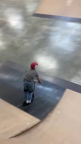 A person wearing a red helmet, gray shirt, and blue jeans with knee pads is riding a scooter inside a skate park. The person is moving from left to right, going up a ramp. Another person is standing in the background observing. The skate park has wooden ramps and structures, with industrial lighting fixtures visible on the ceiling.