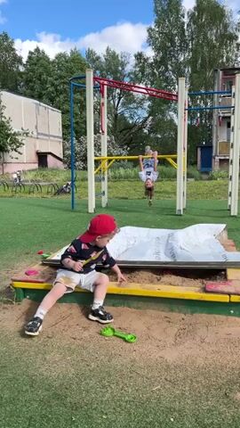 The video depicts a sunny day at a playground. In the foreground, a young child wearing a red baseball cap, a black patterned shirt, and white shorts, is sitting in a sandbox. The child is playing with sand, using a small green toy shovel. Behind the sandbox, a set of monkey bars stands, with a child hanging upside down, gripping the bar with their legs. The playground is covered in artificial green turf. In the background, there are several nondescript, older buildings, trees, and parked bicycles. The sky is blue with scattered clouds. The sandbox is partially covered by a white sheet of plastic.