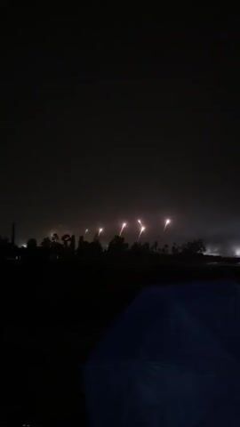A nighttime scene shows a dark sky with distant fireworks. The foreground features the top of a dark blue umbrella. Beyond the umbrella, a silhouette of trees is visible against the faint glow of the fireworks. Smoke or fog drifts across the landscape.
