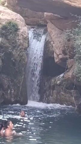 The video shows a serene natural setting with a waterfall cascading into a clear pool of water. Several people are visible in the water, some swimming and others standing. The environment is characterized by large rock formations, lush vegetation, and the sound of flowing water. The overall atmosphere appears relaxed and recreational.