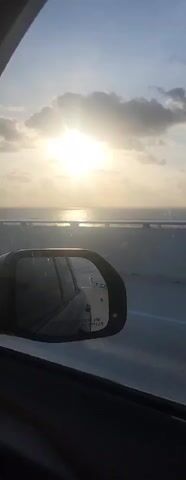 The video shows a view from a car traveling on a bridge over water. The sun is shining brightly, partially obscured by clouds. The ocean stretches out to the horizon, with sunlight glinting on the water. A car's side mirror is visible in the foreground, reflecting the interior of the car and the road behind. The bridge appears to be a multi-lane highway with a barrier separating the lanes. In the distance, a pole with a small sail-like structure is visible, likely part of the bridge's infrastructure. The sky is a pale blue with some white clouds. The overall scene is one of travel and a scenic view of the ocean during daytime.