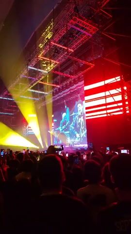 The video shows a concert from the audience's perspective. A large screen displays a band playing, with a drummer on the left and a guitarist/singer on the right. The stage is lit with various colors, including red, yellow, and blue. A complex grid of lights hangs overhead. The audience, visible in the foreground, holds up their phones to record the performance.