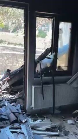 The video shows the interior of a room heavily damaged by fire. The room is filled with charred debris, including wood, brick, and other materials. The walls are blackened with soot, and the paint is peeling. There are multiple windows, some broken and some intact, that offer a view of the outside, which includes a stone wall, some greenery, and a glimpse of other buildings. The overall impression is one of devastation and destruction. No people are visible in the video.