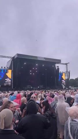 A wide shot captures a large crowd gathered at an outdoor concert venue under a cloudy sky. People, many wearing clear plastic ponchos, are visible in the foreground and extending back towards a large stage complex. The stage is set up with a massive screen and lighting rig. The audience appears to be actively watching the performance. Trees and distant structures are visible beyond the concert area.