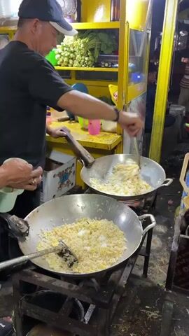 Two people are cooking in a street food stall. The person on the left, wearing a black t-shirt and a black baseball cap, is stirring rice and eggs in a large wok with a spatula. The person on the right, with their arm visible, is also stirring food in a wok with a spatula. The woks are on a portable gas stove. The stall has a yellow exterior with shelves displaying vegetables and food items. The scene appears to be at night, illuminated by artificial lights.