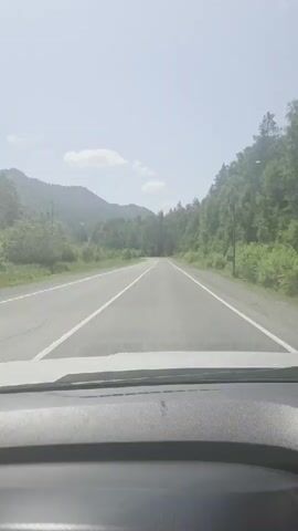 The video shows a view from the inside of a car looking forward through the windshield. A two-lane asphalt road with white lane dividers stretches ahead into a curve. On either side of the road are trees and hills. The sky is bright and hazy, suggesting it is daytime.