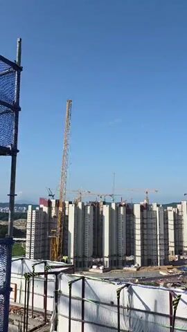 A wide-angle aerial view shows a large construction site under a clear blue sky. Several tall, unfinished apartment buildings are visible, many of them in various stages of completion, with concrete structures and scaffolding. Multiple yellow tower cranes are positioned around the site, some with their booms extending high into the air, actively participating in the construction process. The foreground is partially obscured by a temporary barrier made of white tarpaulin and metal fencing, suggesting a viewpoint from an elevated position on an adjacent structure or platform. In the background, lush green hills and more distant buildings can be seen, indicating an urban or suburban setting. The overall impression is one of ongoing development and large-scale construction activity.