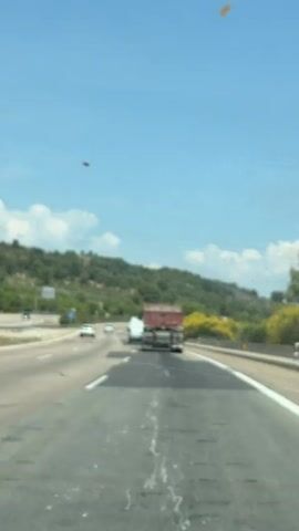 The video shows a car driving on a highway, following a large red truck. The highway is straight and has multiple lanes. On the left side of the road, there are signs and a green hillside in the background. On the right side, there is some yellow vegetation. The sky is blue with scattered clouds. The view is from inside the car, showing the dashboard and a slightly dirty windshield. The car seems to be maintaining a steady speed, with only minor steering adjustments.