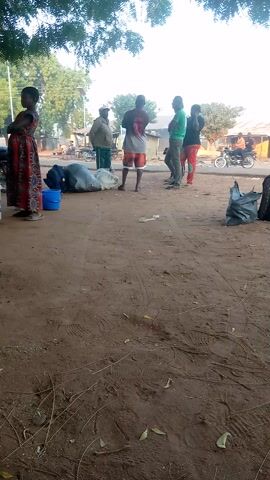The scene shows a group of people standing on a dirt road in a rural area. There are at least six people visible, including a woman in a red and black patterned dress standing in the foreground and five men standing in a line further down the road. The men are wearing various casual clothing. Several bags and a blue bucket are placed near the men on the ground. In the background, there are buildings, trees, and a motorcycle. The road is made of dirt and has some scattered leaves and debris. The overall lighting suggests it is daytime, and there is a tree on the left side of the frame, partially obscuring the view.