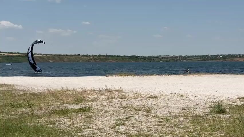 The video shows a body of water with a sandy beach in the foreground and grassy vegetation. In the background, there is a long stretch of land with trees and buildings. The sky is blue with scattered white clouds. There are two kitesurfers on the water, one closer to the shore and one further out. The kitesurfer closer to the shore is in the air, performing a trick. The other kitesurfer is on the water, appearing to be in the process of launching or maneuvering. A person can be seen in the water behind the closer kitesurfer, possibly also participating in watersports. The overall scene depicts a recreational activity on a sunny day at a lake or bay.