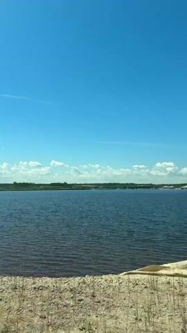 The video shows a large, light-colored rock in the foreground, with a recessed area on top filled with smaller rocks and soil. Behind the rock, a wide body of dark water stretches out under a clear blue sky dotted with a few white clouds. The distant shoreline is lined with trees and what appear to be sandy mounds. The ground around the rock is sandy and uneven, with some sparse vegetation. The camera moves slightly, giving a dynamic view of the scene.