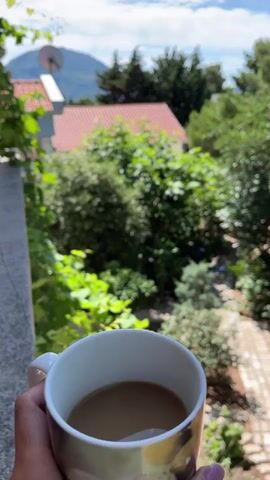 A hand holds a white mug filled with coffee while looking out at a scenic view of trees, a house with a red roof, and a mountain in the distance. The camera pans to the right, revealing more lush greenery and the blue ocean under a clear sky. Sunlight illuminates the scene, creating a tranquil and inviting atmosphere.