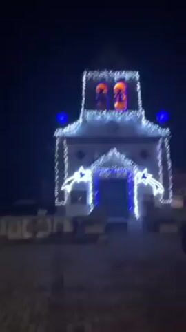 At night, the video opens with a shot of a white church illuminated with white and blue Christmas lights. Two figures stand in the bell tower. The camera pans right, revealing a man walking, then focuses on a large bonfire burning in the middle of a street. People stand nearby.