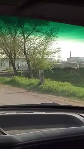 The video is filmed from inside a moving vehicle, presumably a car, focusing on the view through the windshield. The camera moves towards a large, white, industrial-looking building with a series of arched windows or openings. The building is surrounded by sparse greenery and young trees. In front of the building, there is a sign that reads "SIR DARYO INNOVATSIYON TUMANI". Power lines and pylons are visible in the background. The windshield has a greenish tint at the top.