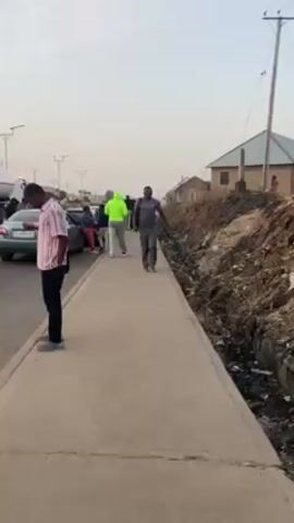 The video shows a man walking on a sidewalk. He is wearing glasses, a gray T-shirt, and gray pants, and he is carrying a black backpack. As he walks closer to the camera, he smiles and spreads his arms wide. Behind him, there is a road with cars, a group of people standing by the cars, and a man wearing a bright green hoodie walking away from the camera. On the other side of the sidewalk, there is a ditch filled with trash and some small buildings. The sky is overcast.