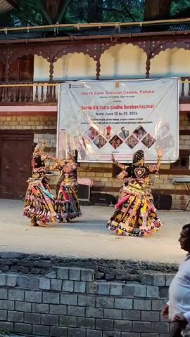 Four female dancers in traditional ornate Indian costumes perform on an outdoor stage. The costumes are brightly colored with intricate embroidery and embellishments, featuring full skirts and elaborate headwear. They are positioned on a stage with a brick facade in the foreground, suggesting an audience view from a lower level. Behind the dancers, a large banner is displayed, indicating an event titled "Sanskriti Yatra-Sindhu Darshan Festival" organized by the North Zone Cultural Centre, Patiala. The backdrop appears to be an open-air venue with architectural elements suggesting a traditional Indian aesthetic, possibly with trees visible above. Two male figures, one adult and one child, are partially visible in the foreground, observing the performance. The lighting suggests it is late afternoon or early evening.