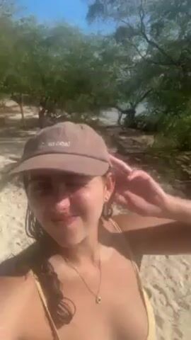 A woman in a bikini and cap is recording a video on a beach. The beach appears to be sandy, with trees and shrubs in the background. The water is visible in the background, reflecting the sunlight. The cap is brown with the word "Charcoal" embroidered on it. The woman looks into the camera and says something. The camera is unsteady, moving slightly while recording.
