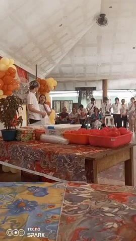 The video shows an indoor event with a stage area decorated with orange and yellow balloons. Two young people, a male and a female, are standing at a table on the stage, seemingly interacting with each other. The female is holding a microphone. In front of them on the table are various items, including what appear to be canned goods, a potted plant, plastic cups, and two large red containers filled with tomatoes. Behind the stage, an audience is seated, observing the event. Several other young people, possibly participants or staff, are standing in the background. The environment appears to be a covered outdoor or semi-outdoor space with a visible ceiling structure.