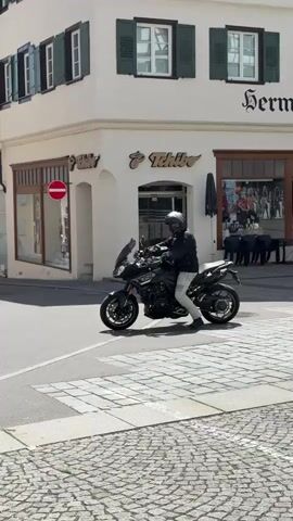 The video shows two motorcycles driving on a paved street in front of a white building with green shutters. The first motorcycle is black and gray and the rider is wearing a black jacket, gray pants, and a full helmet. The second motorcycle is black and silver and the rider is wearing black attire and a full helmet. The building behind them has a sign that says "Hermann Dorner" and another sign that says "Tchibo." There is a "Do Not Enter" sign on the corner of the building. In the background, two people are walking. The street is paved with cobblestones. The day is sunny.