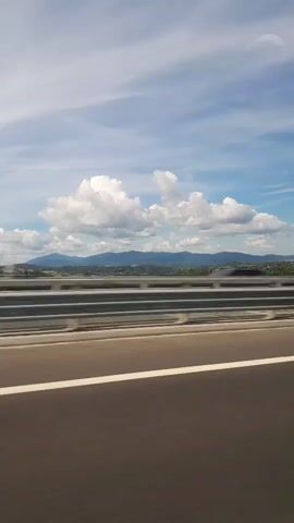 A video taken from a moving vehicle shows a view of a highway. In the foreground is the asphalt road with a white line. A metal guardrail separates the road from the opposite lanes. Beyond the guardrail, a landscape is visible with mountains in the distance under a blue sky with scattered clouds. A few cars can be seen on the opposite side of the highway. The video is shaky, indicating movement.