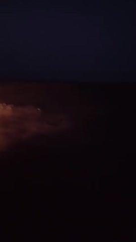The video displays a dark sky with subtle hues of orange and red near the horizon, indicating either dusk or dawn. The lower portion of the frame is dominated by a deep shadow, obscuring any discernible details of the ground or objects. There are no visible people, actions, or interactions. The environment appears to be outdoors, possibly a landscape or seascape, with the darkness suggesting limited light conditions. The overall impression is one of a quiet, almost abstract scene of the sky during twilight.