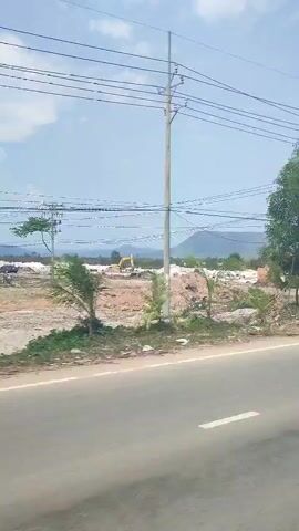 The video shows a view from a moving vehicle driving on a paved road. In the first shot, an excavator is visible in the background, in a construction area.  There are power lines overhead. In the second shot, a scooter with two people riding it crosses the road at a pedestrian crossing. In the third shot, the vehicle passes by a white shack, with equipment and a covered stall in front of it. There are trees around the shack.