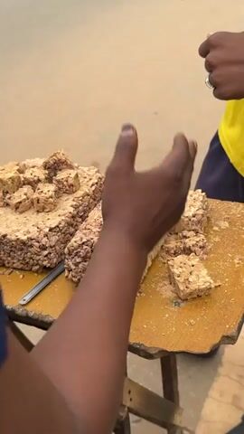 A first-person shot shows a table with large slabs of peanut brittle. The brittle has been cut into smaller squares. A hand enters the frame and hovers over the brittle, seemingly measuring the size. Another person in a yellow shirt stands to the right, presumably the vendor. The table is wooden and sits on a wooden stand. The background is blurred, suggesting an outdoor setting.