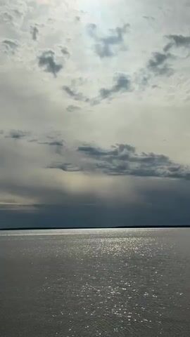 The video shows a wide expanse of water under a cloudy sky. The sun is visible behind the clouds, creating a silvery, shimmering effect on the water's surface. The horizon line is visible in the distance, with a dark strip of land barely discernible. No people or objects are visible in the scene other than the water and the sky. The camera appears to be stationary.