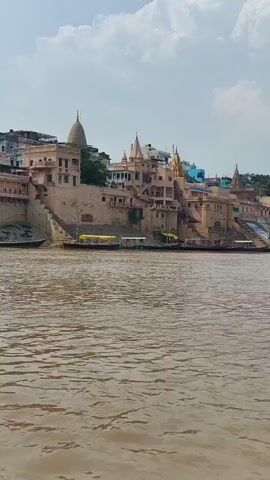 The video shows a wide view of the Ganges River with buildings and ghats on the opposite bank. Several boats are docked along the river's edge. The buildings appear to be a mix of religious structures and residences, with a prominent temple spire visible in the distance. The water is a murky brown color. The sky is partly cloudy.