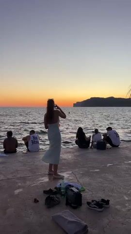 A group of six young people are sitting on a concrete pier watching the sunset over the ocean. The sky is a gradient of orange and purple. The ocean is calm with small waves. In the foreground, there are various items on the ground including shoes, a bag, a towel, and a bottle of water. A woman in a light blue dress stands in the center of the frame, facing away from the camera, with her hair blowing in the wind. The other five people are seated on the edge of the pier, facing the ocean.