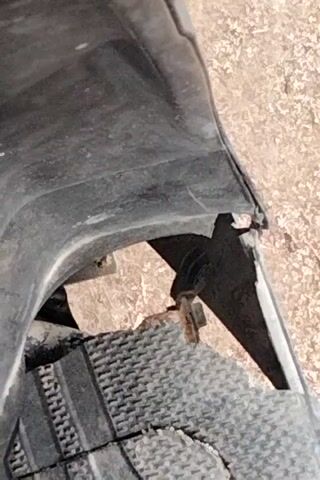 The video shows a close-up of a damaged black plastic bumper on a vehicle. The bumper is torn and broken, with a piece of grey material with a textured pattern protruding from the tear. A rusty bolt is visible within the damaged area. The background appears to be a dusty or gravelly surface. The camera moves slightly, offering different angles of the damage.