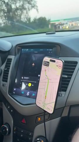 The video shows the interior of a car. A smartphone is mounted on the dashboard, displaying a navigation app. The screen of the car's infotainment system is also visible, showing music playback controls and the song title "Amsterdam". The car appears to be moving, as indicated by the blurred background and the navigation app showing a route. A gas station is visible in the background.